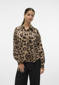 Blusa in satin beige e marrone con stampa leopardata, dotata di colletto, chiusura frontale con bottoni e maniche lunghe. Indossata con pantaloni neri.