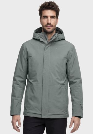 Mann mit kurzen Haaren und Bart, der eine grüne Kapuzen-Winterjacke und schwarze Hose trägt, steht vor einem einfarbigen hellen Hintergrund.