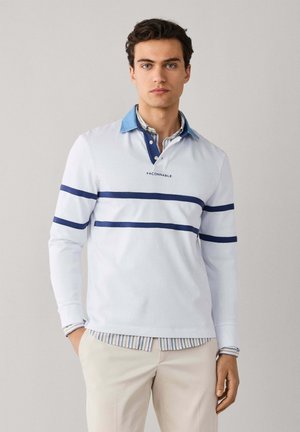 Jeune homme portant un maillot de rugby blanc à manches longues avec des rayures horizontales bleues et un col bleu, associé à un pantalon beige clair.
