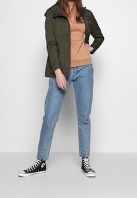 Oliwkowo-zielona kurtka z kapturem, beżowy sweter i jasnoniebieskie dżinsy. Czarne wysokie trampki z białymi podeszwami. Codzienny, warstwowy strój.