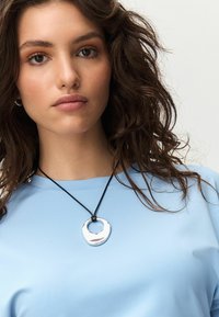 Sølvfarvet metalvedhæng på en sort snor, der har en uregelmæssig cirkulær form. Bæreres over en lyseblå, kortærmet top.