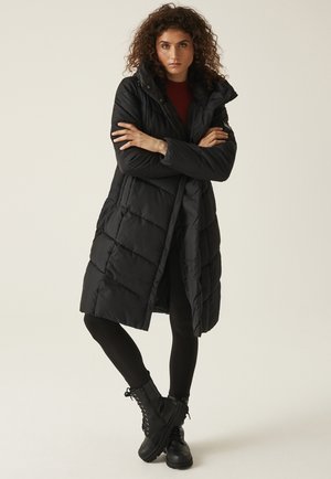 Kobieta z kręconymi włosami, ubrana w długą czarną pikowaną kurtkę, czarne legginsy i czarne sznurowane botki, stojąca z założonymi ramionami na jednolitym tle.