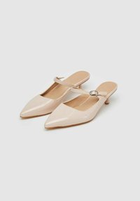 Talons aiguilles à bride arrière en cuir verni nude, avec un bout pointu, une fine sangle de cheville et un petit talon bloc. Détail de la semelle texturée visible.
