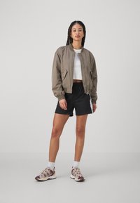 Chaqueta bomber marrón, top corto blanco, pantalones cortos deportivos negros con rayas laterales y zapatillas marrón/crema. Cabello trenzado, fondo neutro.