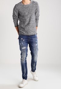 Šedý textured svetr, světle modré džínové kalhoty s efektivním opotřebením a bílé tenisky. Oblečen í má volný střih a kulatý výstřih.