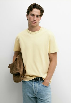 Junger Mann mit welligem Haar trägt ein hellgelbes T-Shirt und hellblaue Jeans, hält eine gefaltete braune Jacke in einer Hand.