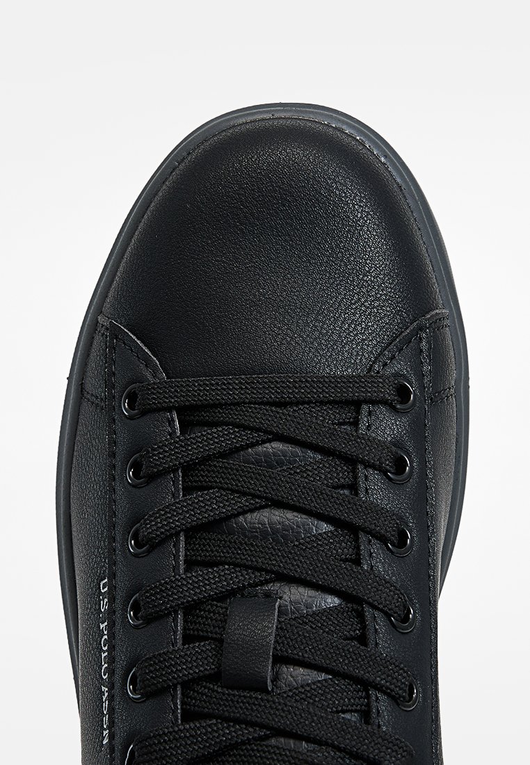 Zapatillas de cuero negro con puntera redondeada, acabado texturizado, cordones planos negros y suela de goma en contraste. Marca U.S. Polo Assn. visible.