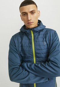Giacca isolante blu con design trapuntato, zip gialla e cappuccio. Tessuto elastico sulle maniche e nei pannelli laterali, logo sul petto.
