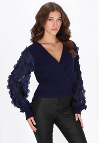 Laivastonsininen kietaisuneule, jossa on teksturoidut, kuusikulmakuviolliset läpikuultavat hihat. Mallissa on resorikuvioitu neulosmateriaali ja syvä V-kaula-aukko. Yhdistetty mustiin housuihin.