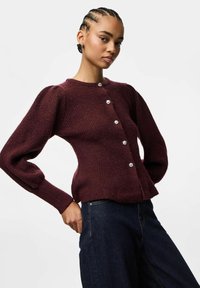 Bordo adīts kardigāns ar pūkām piedurknēm, apaļu kakla izgriezumu un pieciem dekoratīviem pogām. Mīksta tekstūra ar pieguļošu siluetu.