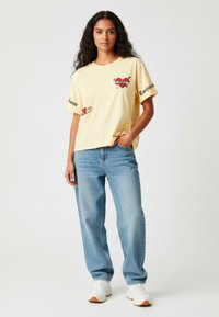 Geel T-shirt met korte mouwen met verschillende kleurrijke bedrukte ontwerpen, gecombineerd met lichtblauwe losse denim jeans en witte sneakers.