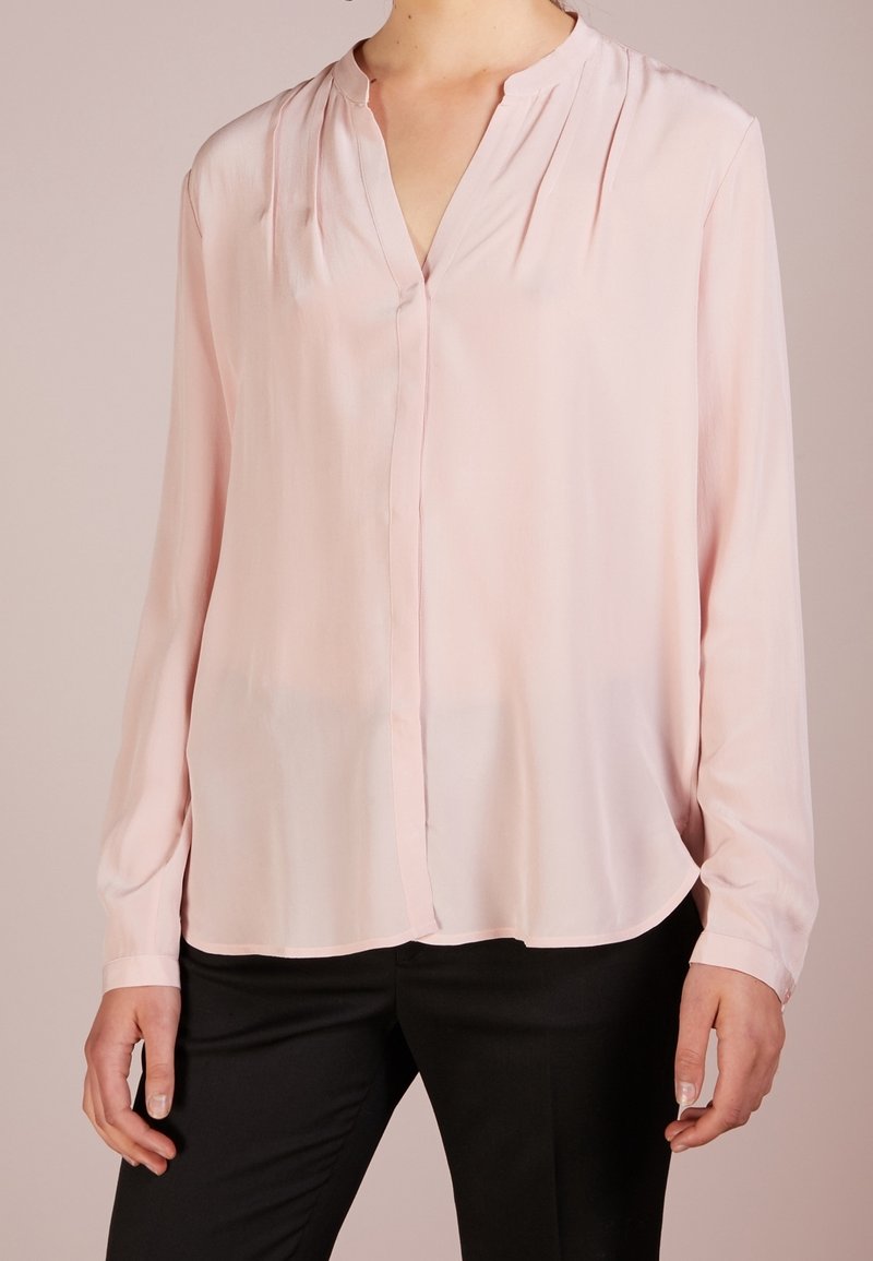 Femme portant un chemisier rose clair à manches longues avec un col en V et un pantalon noir, debout devant un fond uni.