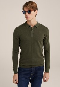 Sötétzöld hosszú ujjú pólóing, három gombos hónaljjal és bordázott mandzsettákkal. A modellen barna keretes napszemüveg és kék farmer van.