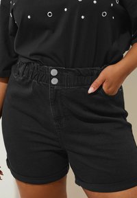 Mustat farkkushortsit, joissa on rypistetty vyötärö ja kaksi hopeista nappia sekä käännetyt lahkeet. Suora pinta, jossa on vähäisiä yksityiskohtia.