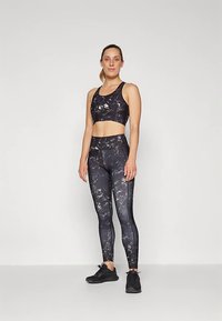 Brassière de sport noire à motifs et leggings assortis, fabriqués en tissu extensible avec un subtil motif floral, associés à des chaussures de sport noires.