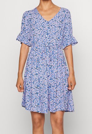 Kobieta ubrana w niebieską sukienkę w kwiaty do kolan, z krótkimi, falowanymi rękawami i paskiem wiązanym w talii.