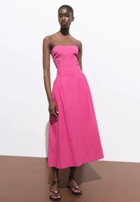 Abito senza spalline a linea ad A di un vivace colore rosa, con dettagli arricciati sul busto e un tessuto fluido che arriva a metà polpaccio. Si indossano sandali neri.