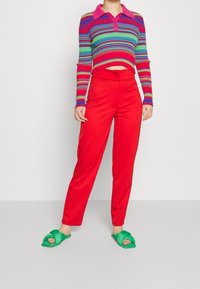 Personne portant un pantalon taille haute rouge, un polo court à manches longues rayé multicolore et des sandales ouvertes vertes, se tenant devant un mur blanc.