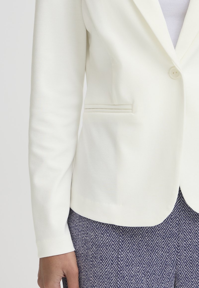 Gros plan d'une personne portant un blazer blanc à un seul bouton et un pantalon bleu marine avec un motif en chevrons.