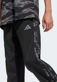 Musta urheiluhousut, joissa on naamiointikuosiset sivu raidat ja harmaalla Adidas-logolla. Valmistettu pehmeästä kankaasta, jossa on rento istuvuus.
