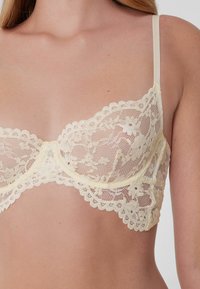 Krajková podprsenka v jemné krémové barvě s vlnitými okraji, zdobená květinovými vzory. Nastavitelné ramínka a kosticová podpora. Jemná textura.