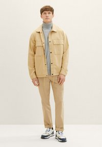 Veste beige clair sur un col roulé gris, associée à un pantalon beige et des baskets multicolores. Présente deux poches poitrine et un ourlet à cordon.