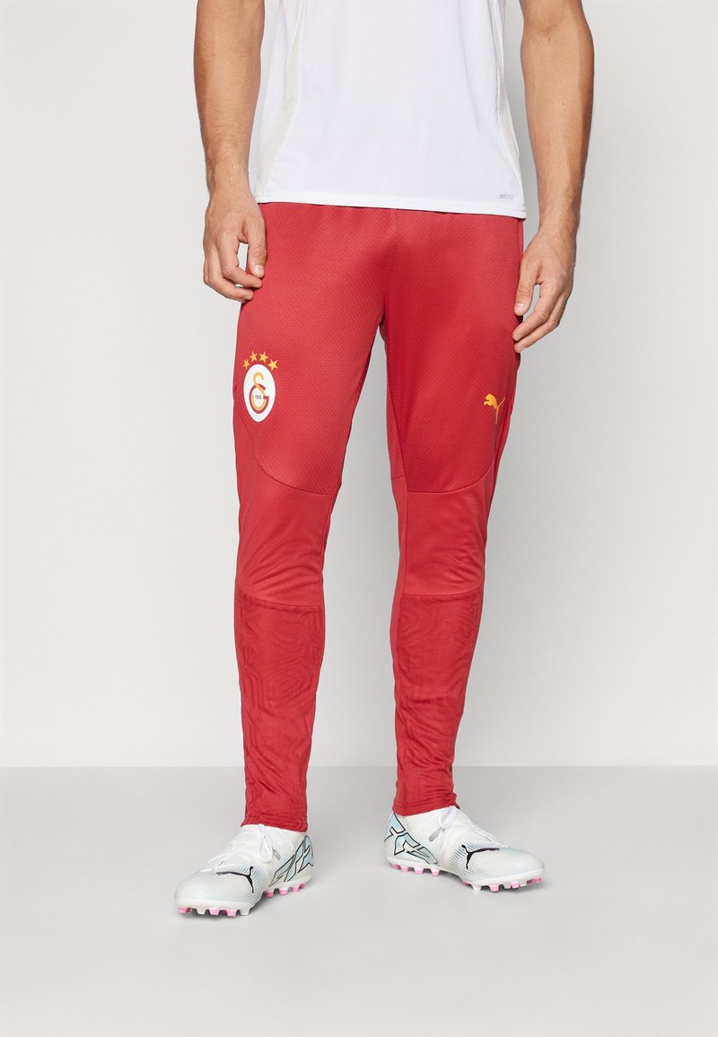 Pantalon de sport rouge avec une finition texturée, présentant un logo jaune sur la cuisse et une taille élastique, assorti de crampons blancs.