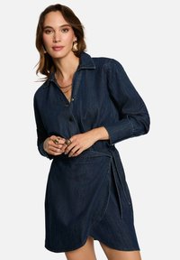 Robe en denim à manches longues, col et fermeture à boutons. Présente une ceinture à la taille et un ourlet légèrement incliné, de couleur bleu indigo foncé.