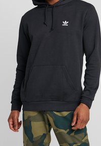 Black hoodie with a drawstring hood, front pocket, and white Adidas logo. Soft fabric, ribbed cuffs, and hem. Paired with camo-patterned pants.