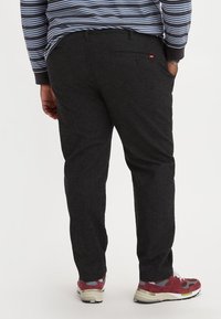 Donkergrijze broek van een gestructureerde stof, met een achterzak en een klein logo-label. Gecombineerd met rood en grijs gemusterde sneakers.