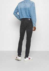 Vaqueros negros ajustados con un detalle desgastado en la pierna, combinados con una camisa de mezclilla azul clara y zapatillas blancas con acentos rojos.