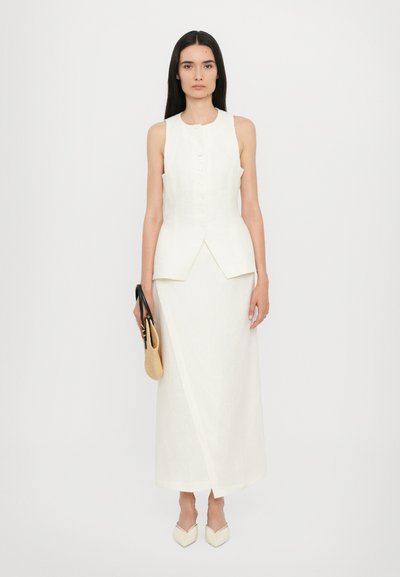 White linen sleeveless top with a V-shaped hem, paired with a matching long skirt. The look is complemented by a black handbag and pointed white shoes.