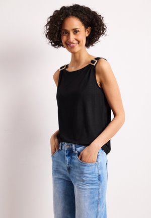 Femme aux cheveux bouclés souriante, portant un haut noir sans manches avec des bretelles à boucle et un jean bleu clair, les mains dans les poches.