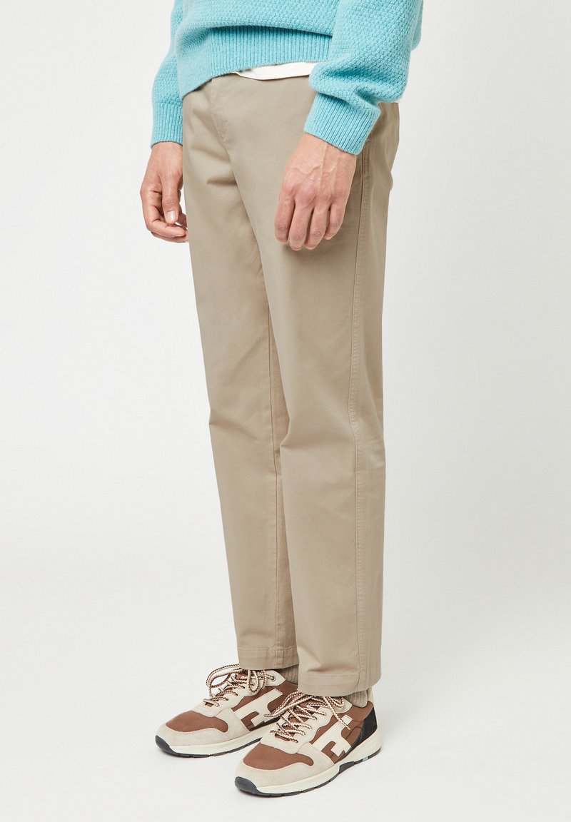 Uomo che indossa pantaloni beige, sneakers marroni e beige e un maglione lavorato a maglia azzurro chiaro, in piedi contro uno sfondo bianco uniforme.
