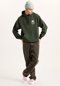 Grøn hættetrøje med frontlomme og logo, kombineret med mørke jeans og lyserøde sneakers. Teksturen ser blød ud, og stofferne har en afslappet og løs pasform.