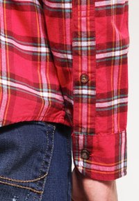 Camicia a quadri rossi con strisce blu e gialle, realizzata in tessuto leggero, con bottoni marroni e un design dei polsini casual.