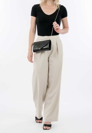 Donna che indossa una maglietta nera a maniche corte e pantaloni beige a gamba larga, con una borsa a spalla nera con tracolla a catena e scarpe nere con tacco aperto.