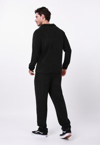 Juodas fliuso sportinis kostiumas su ilgomis rankovėmis ir kelnėmis, turintis laisvą siluetą ir lygią faktūrą. Papildytas juodai baltais sportbačiais.