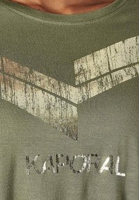 T-shirt vert olive avec logo imprimé en or et argent métallisé ; présente une texture usée et un col rond. Tissu léger.