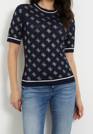 Frau trägt ein marineblaues kurzärmeliges Stricktop mit weißem Logo-Muster und weißer Einfassung, kombiniert mit hellblauen Jeans.