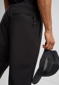Los pantalones atléticos negros presentan un bolsillo trasero con cremallera, una textura suave y una cinturilla elástica. Una gorra oscura con un panel de malla se sostiene en una mano.