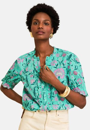 Kvinde med kort krøllet hår iført turkis blomsterbluse og cremefarvede bukser, accessoriseret med guldringe og armbånd, poserer selvsikkert.