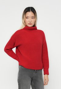 Frau mit langem Ombre-Haar, die einen roten Strickpullover und graue Jeans trägt. Sie steht mit einer Hand auf der Hüfte und schaut seitwärts.