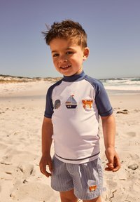 Weißes Rashguard mit kurzen Ärmeln, blauen Akzenten und nautischer Stickerei, kombiniert mit blau-weiß gestreiften Shorts mit einem orangefarbenen Fuchs.