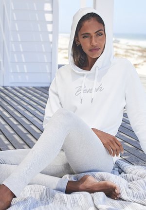 Frau in weißem Hoodie und hellgrauen Leggings sitzt barfuß auf einer gestreiften Decke auf einem Holzdeck am Strand.