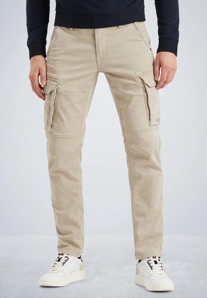 Pantalon cargo beige avec poches à rabat porté avec des baskets blanches et un haut noir à manches longues, montré de la taille aux pieds.