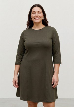 Une femme souriante porte une robe olive foncé jusqu’aux genoux, à manches trois quarts, et des créoles dorées, sur un fond uni et clair.
