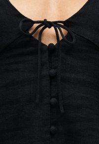 Μαύρο υφασμάτινο τοπ με υφή, με τέσσερα καλυμμένα με ύφασμα κουμπιά στο κέντρο και έναν δεμένο φιόγκο στο ντεκολτέ πάνω από μια μικρή V-σχήματος εγκοπή.