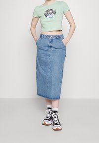 Osoba nosící světle zelené krátké tričko s grafikou cheruba, modrou džínovou midi sukni s vysokým pasem, bíl é ponožky a černo-bílé tenisky.