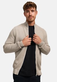 Beige rits-in-sweater met een gestructureerd gebreid patroon. Voorzien van een hoge kraag en ribgebreide manchetten. Gedragen over een zwart T-shirt.